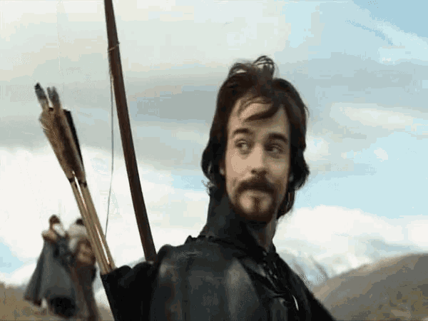 a man with a beard is holding a bow and arrow in a field .