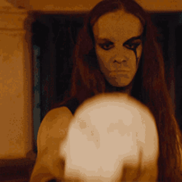 a woman with long hair and white paint on her face is holding a skull