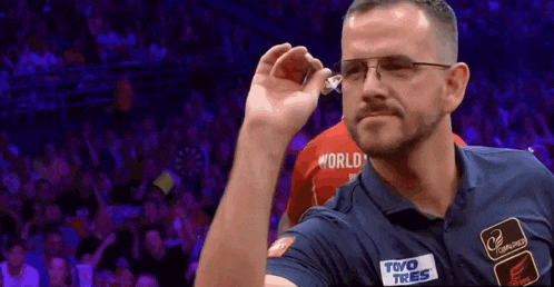 a man wearing glasses and a toyo tires shirt throws a dart at another man