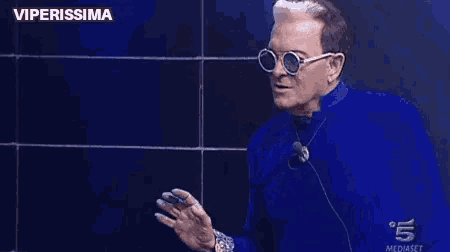 a man wearing sunglasses and a blue jacket is waving his hand .