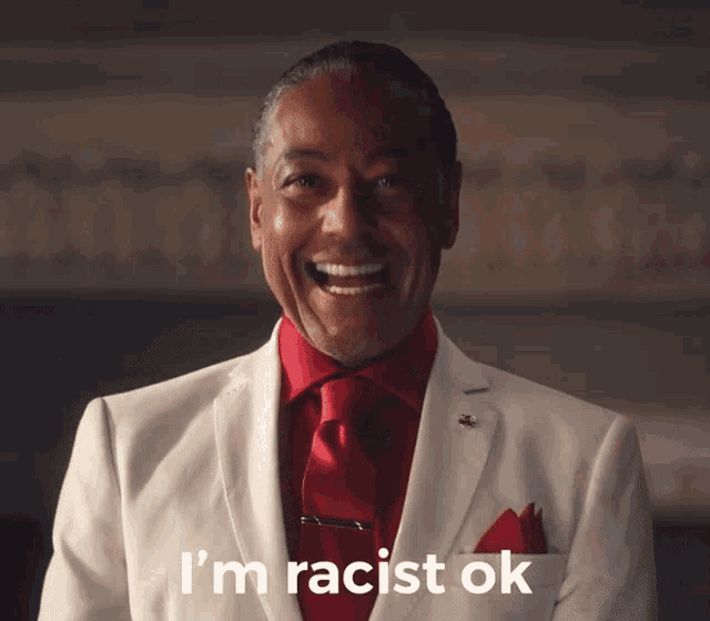 a man in a white suit and red tie is smiling and says i 'm racist ok