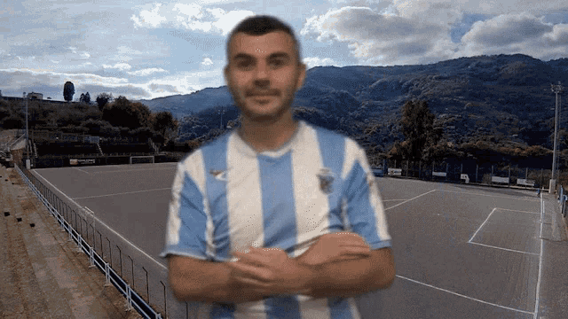 a man in a blue and white striped shirt is standing in front of a soccer field with mountains in the background