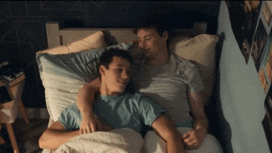 two young men are laying in bed with their arms around each other and one of them is wearing a blue shirt