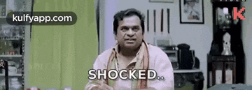 a man in a pink shirt is sitting in a living room and looking shocked .