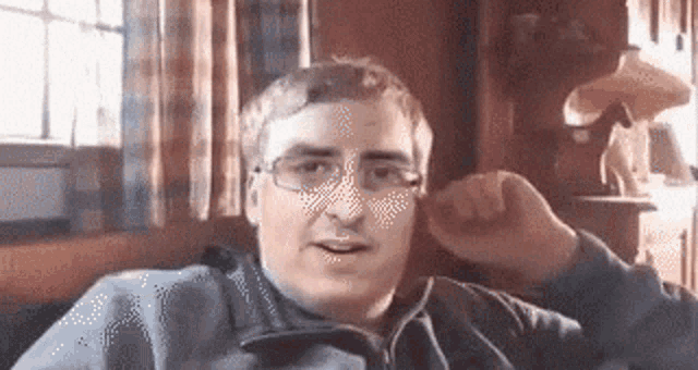 a man wearing glasses is sitting on a couch in front of a window .