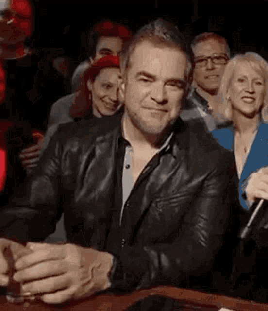 a man in a leather jacket is sitting at a table holding a glass of whiskey in front of a crowd of people .