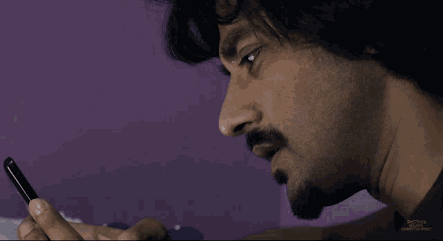 a man with a beard is looking at a cell phone with a purple wall behind him