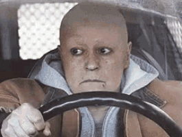 a bald man is driving a car and looking at the camera .