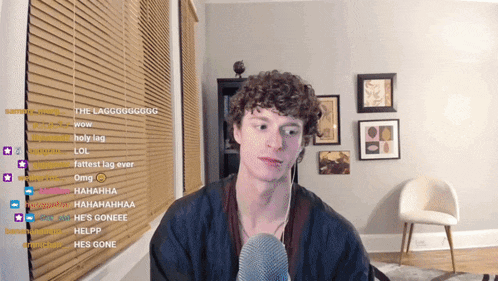 a man with curly hair stands in front of a microphone in front of a window with blinds on it