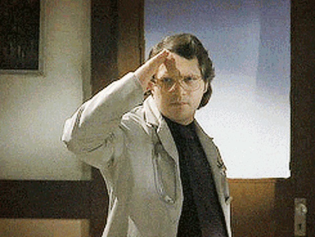 a man wearing glasses and a white jacket salutes while standing in front of a door