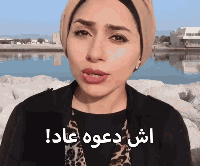 a woman wearing a leopard print scarf stands in front of a body of water with arabic writing on the bottom of her face