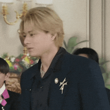 a man in a suit with a brooch on his jacket is standing in a room with other people .