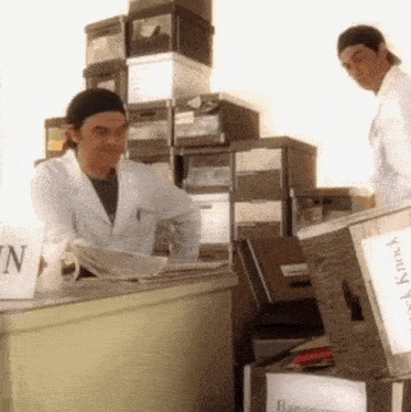 a man in a lab coat sits at a desk in front of a stack of filing cabinets with boxes labeled knock