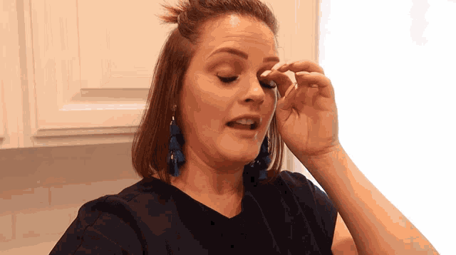 a woman wearing a blue shirt and blue earrings wipes her eye with her hand
