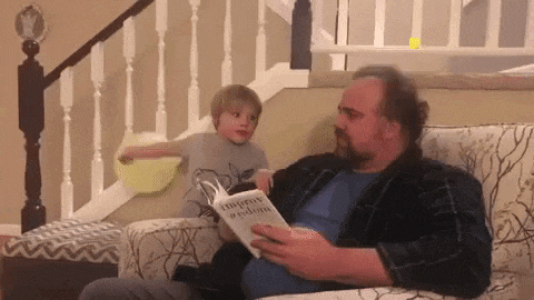 a man is sitting on a couch reading a book to a child .