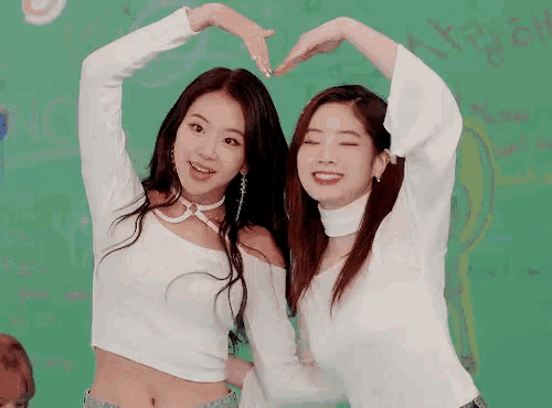 two girls are making a heart shape with their hands in front of a green board .