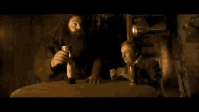 a man with a beard is sitting at a table holding a bottle of wine and a glass .
