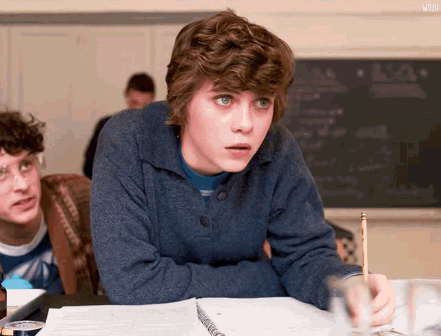 a boy with curly hair sits at a desk with a notebook and a pencil