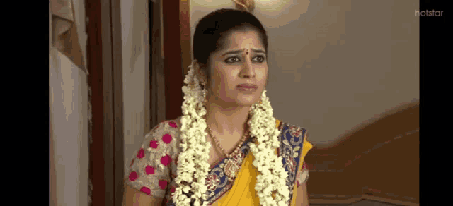 a woman in a yellow saree with a garland of flowers around her neck is sitting in a room .