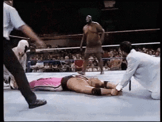 a wrestler is laying on the ground in a wrestling ring while a referee stands behind him .
