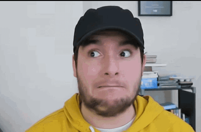 a man with a beard wearing a black hat and a yellow hoodie makes a funny face