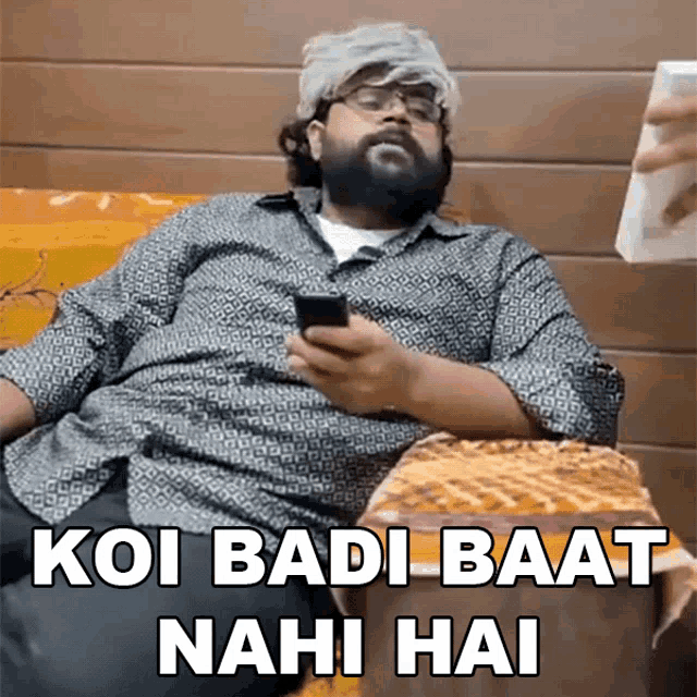 a man with a beard is sitting on a couch holding a cell phone with the caption koi badi baat nahi hai