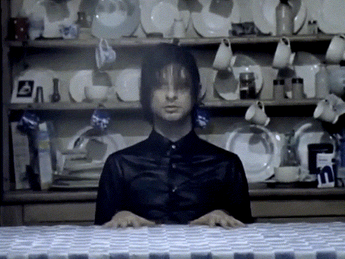 a man in a black shirt is sitting at a table in front of a shelf full of plates and cups
