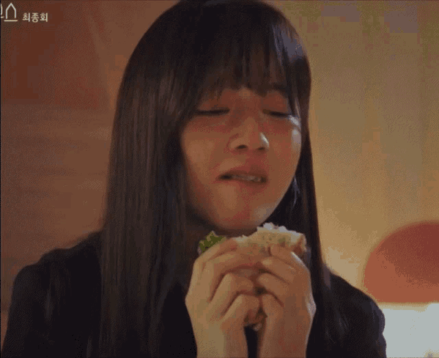 a woman with long hair is eating a sandwich with korean writing on the bottom right