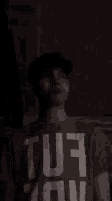a young man wearing a hat and a t-shirt with the word tuf on it is standing in the dark .