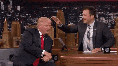 a man in a suit and tie is touching another man 's head while sitting in front of a microphone .