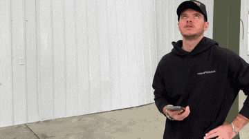 a man wearing a hat and a black hoodie holds a cell phone and looks up