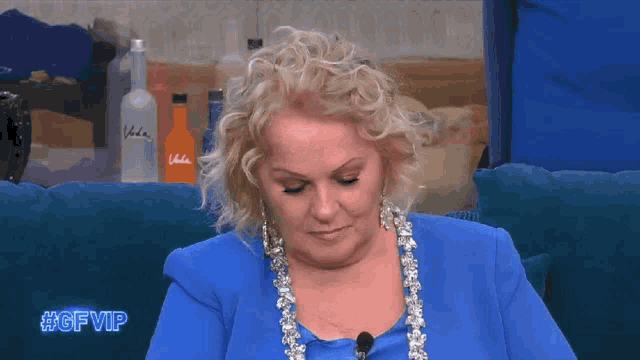 a woman in a blue suit is sitting on a blue couch with a bottle of vodka in the background