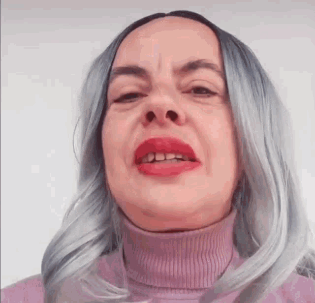 a woman with gray hair and red lipstick is wearing a pink turtleneck sweater and making a funny face .