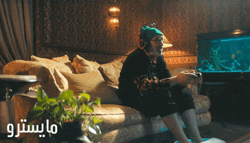 a man sits on a couch playing a video game with arabic writing behind him