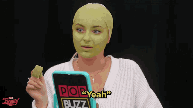 a woman with a green mask on her face is holding a phone that says pop yeah buzz