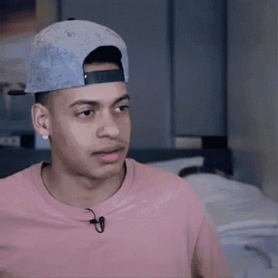 a young man wearing a baseball cap and a pink shirt is sitting in a bedroom .