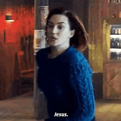 a woman in a blue sweater is standing in a room and saying `` jesus '' .