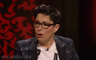 a woman wearing glasses and a white t-shirt is sitting in front of a screen that says #taskmaster