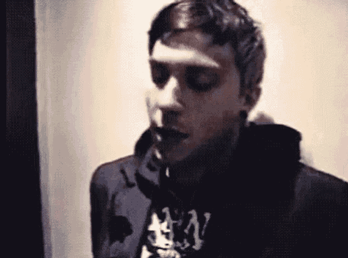 a man in a black hoodie is smoking a cigarette while standing in front of a wall .