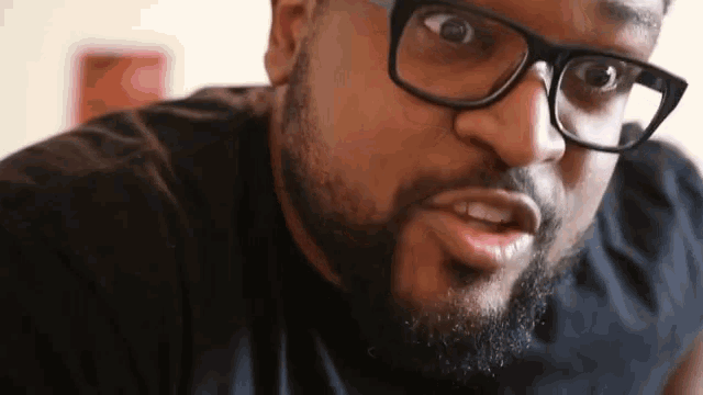 a man with a beard wearing glasses and a black shirt is making a funny face .