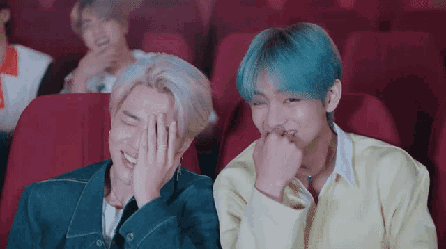 a man with blue hair is covering his face while sitting next to another man