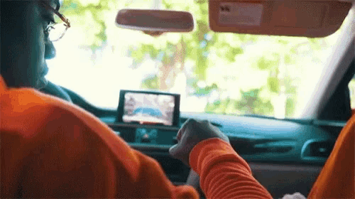 a man in an orange shirt is driving a car with a rear view camera on the dashboard .