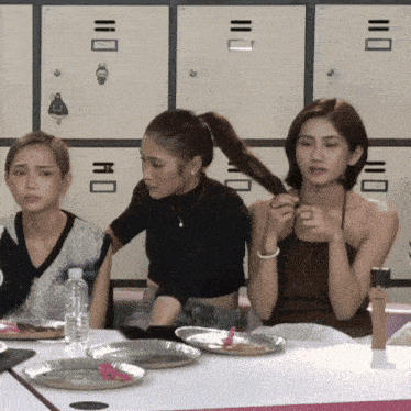 three women are sitting at a table with plates and a bottle of water in front of lockers with the numbers 1 through 10 on them