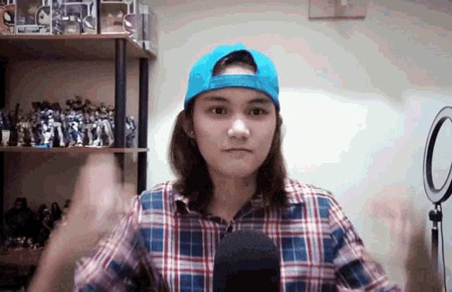 a woman wearing a plaid shirt and a blue hat stands in front of a shelf full of action figures