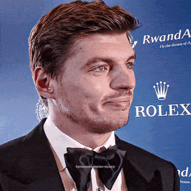 a man in a tuxedo and bow tie stands in front of a rolex sign