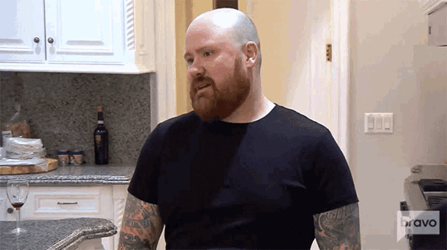 a bald man with a beard is standing in a kitchen wearing a black shirt that says bravo on the front