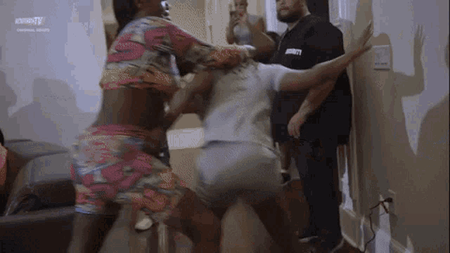 two women are fighting in a living room with a man in a security guard watching .