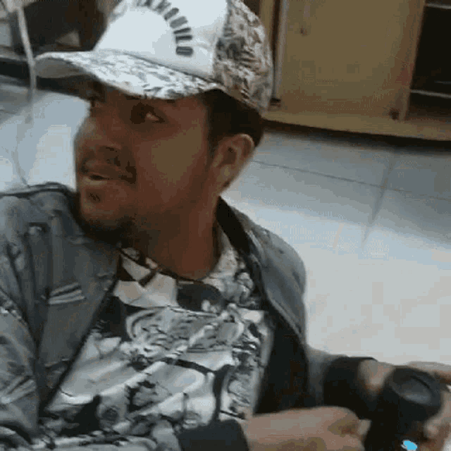 a man wearing a hat and a jacket is sitting on the floor holding a remote control .