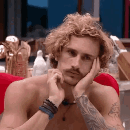 a shirtless man with curly blonde hair and a tattoo on his arm is sitting on a red chair with his hands on his face .