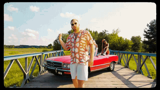 a man in a colorful shirt is standing on a bridge next to a red mercedes